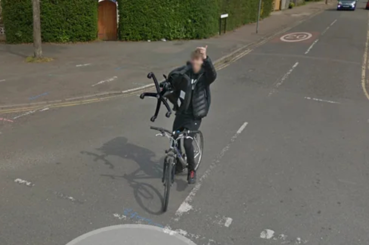 Person on a bike waving at the camera with one hand, obscuring their face, on a suburban street