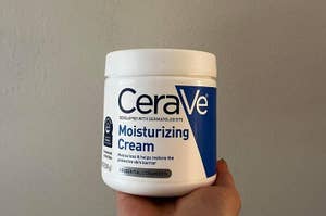 Hand holding a CeraVe Moisturizing Cream container against a plain background
