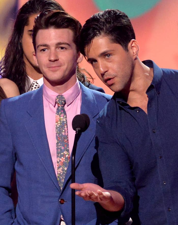 Adult Drake Bell and Josh Peck standing onstage as Josh speaks into a microphone