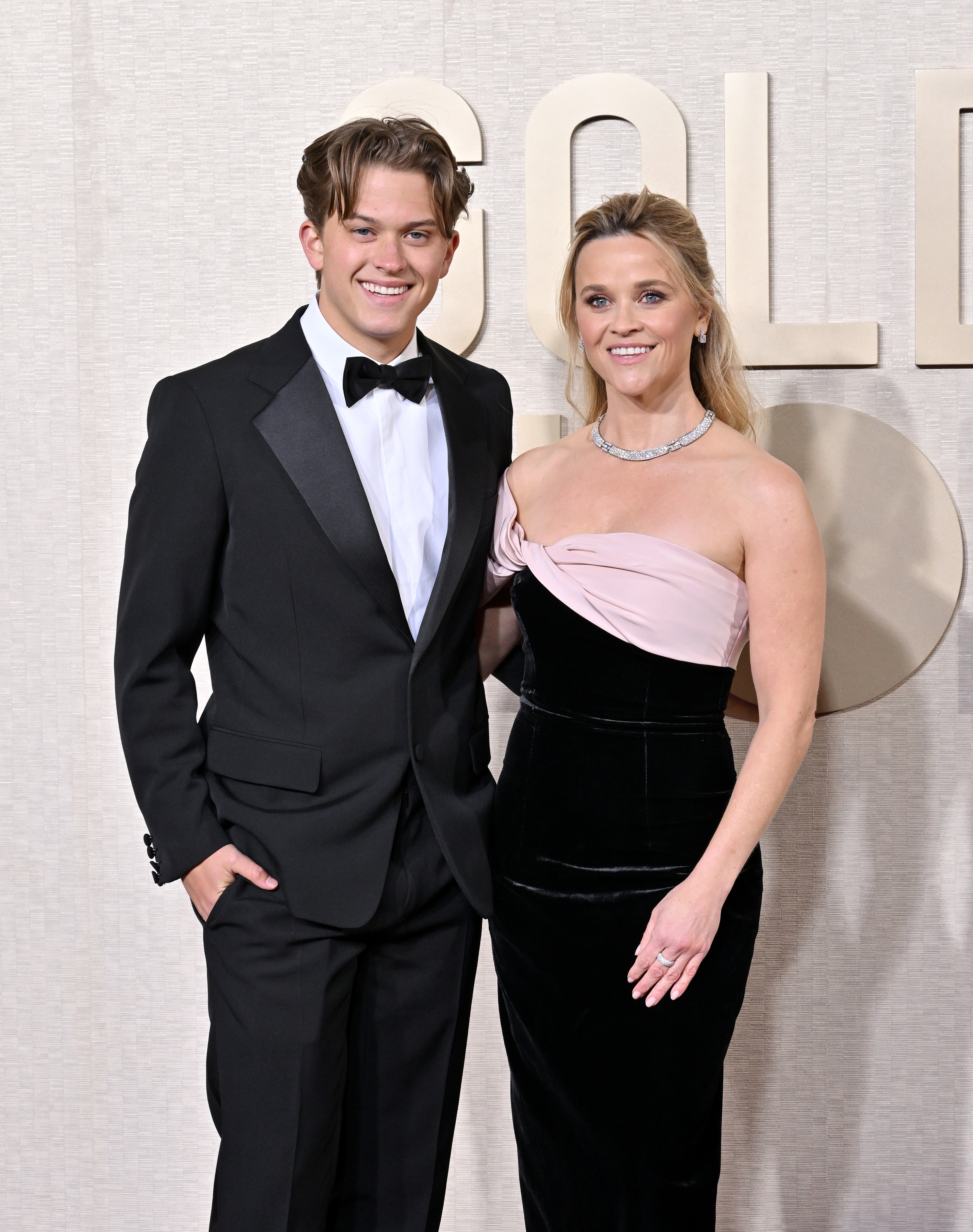 Deacon in a black tuxedo and Reese in a strapless gown, both smiling, at a media event
