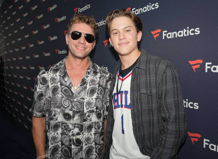 Ryan and his son, Deacon, posing together at a media event, Ryan in a floral shirt and sunglasses, the other in a plaid jacket with a graphic T-shirt