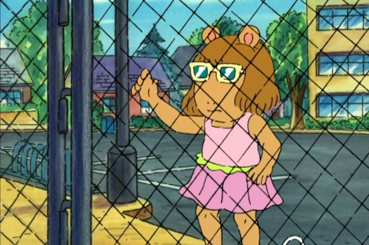 Arthur character D.W. stands by a fence with one arm raised, wearing glasses and a pink dress