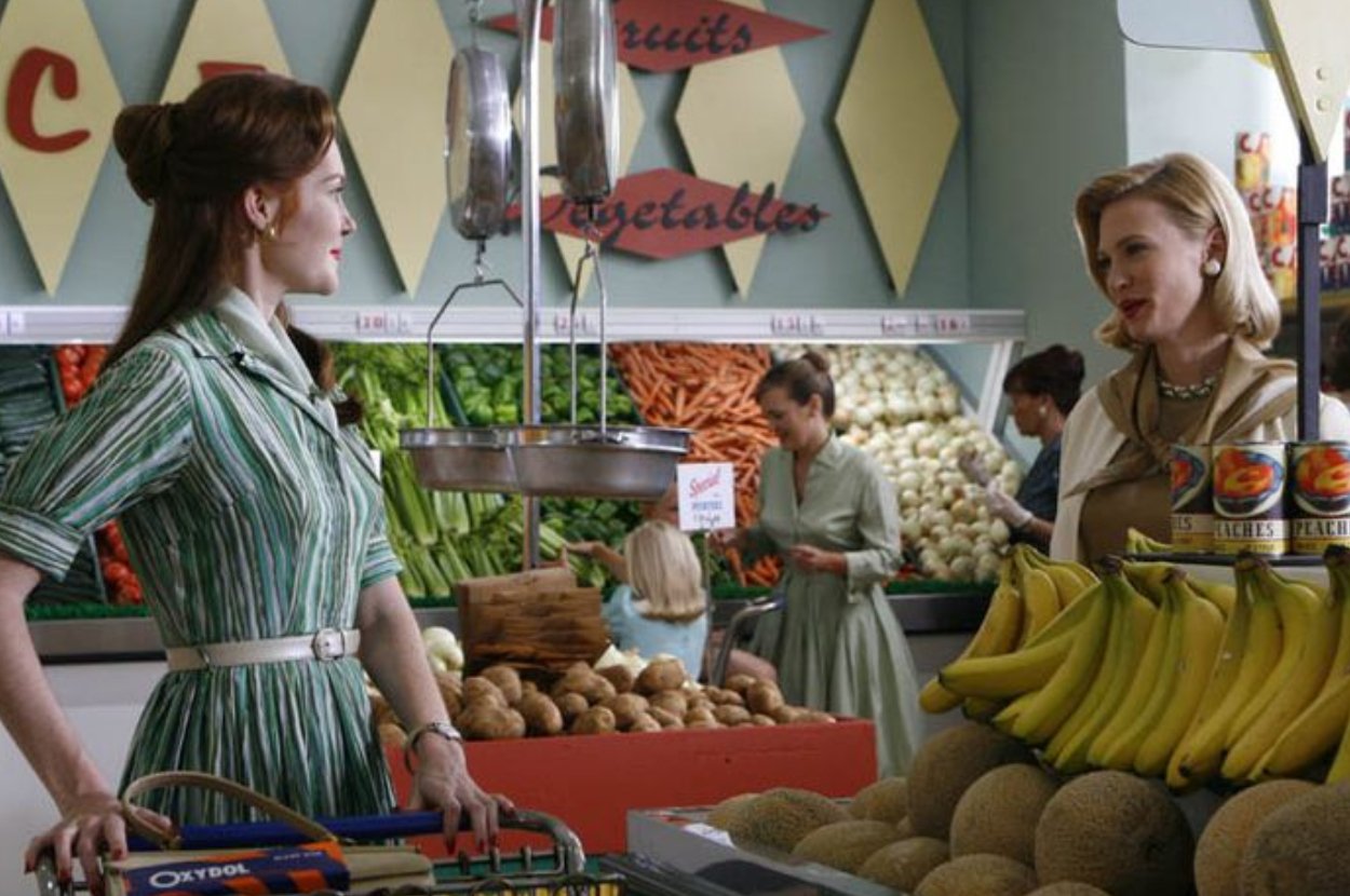 Two actresses in a vintage grocery store scene, dressed in period costumes with a shopping cart