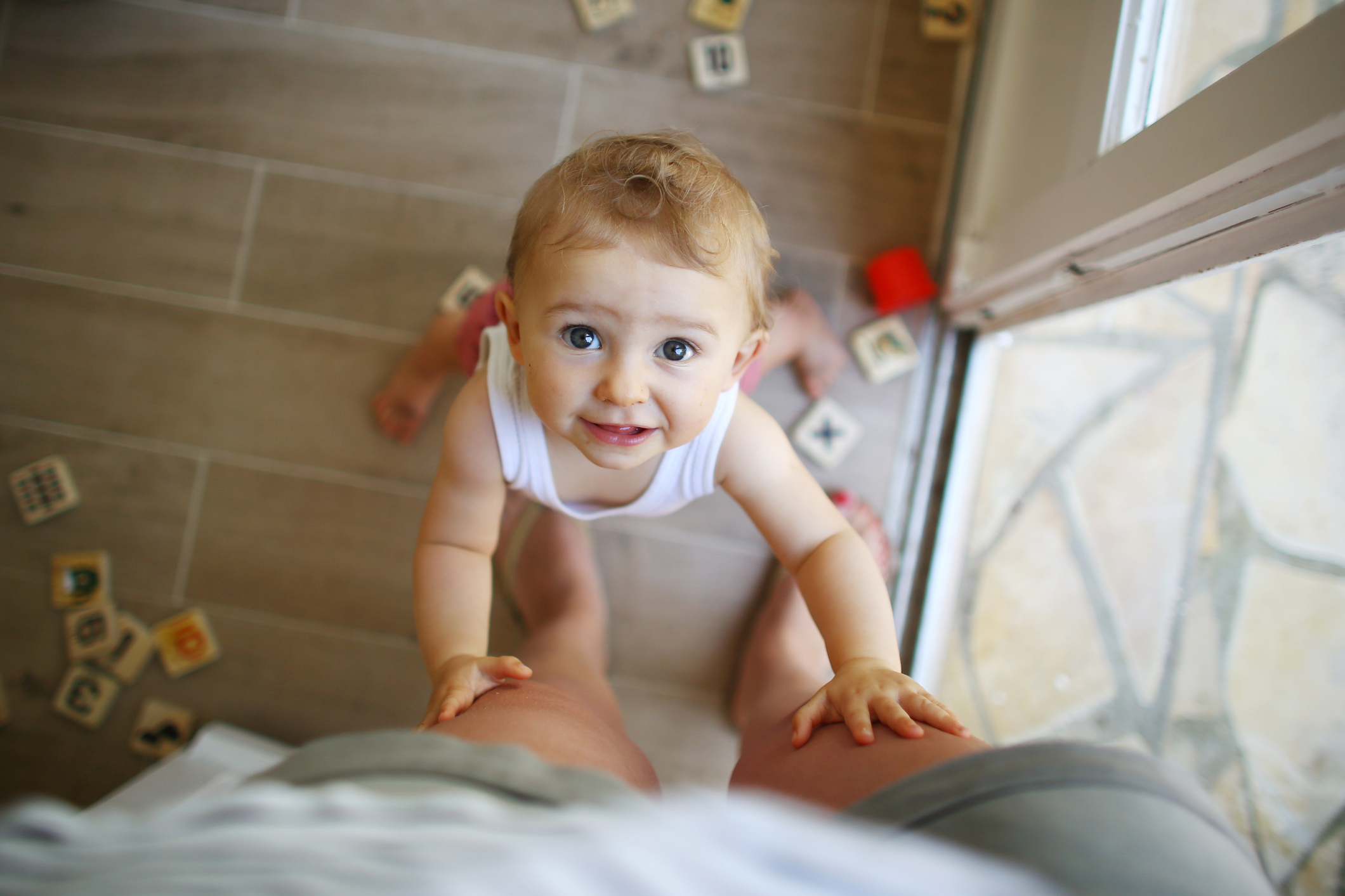 Baby climbing onto adult&#x27;s knee, looking up at the camera, alphabet blocks scattered around on the floor