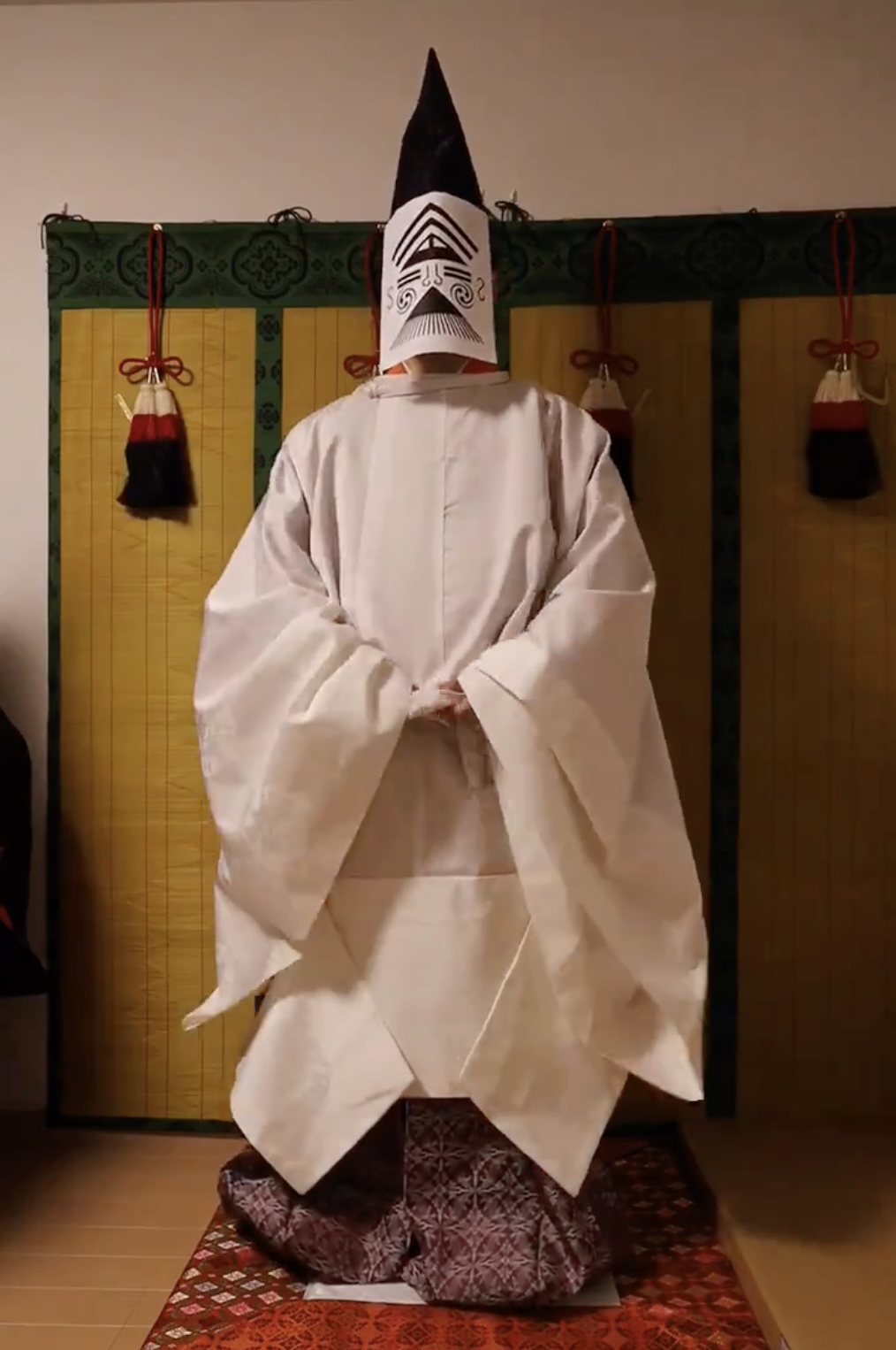 伝統的な装束を身にまとった能楽の演者が、和室で背後から撮影されています。