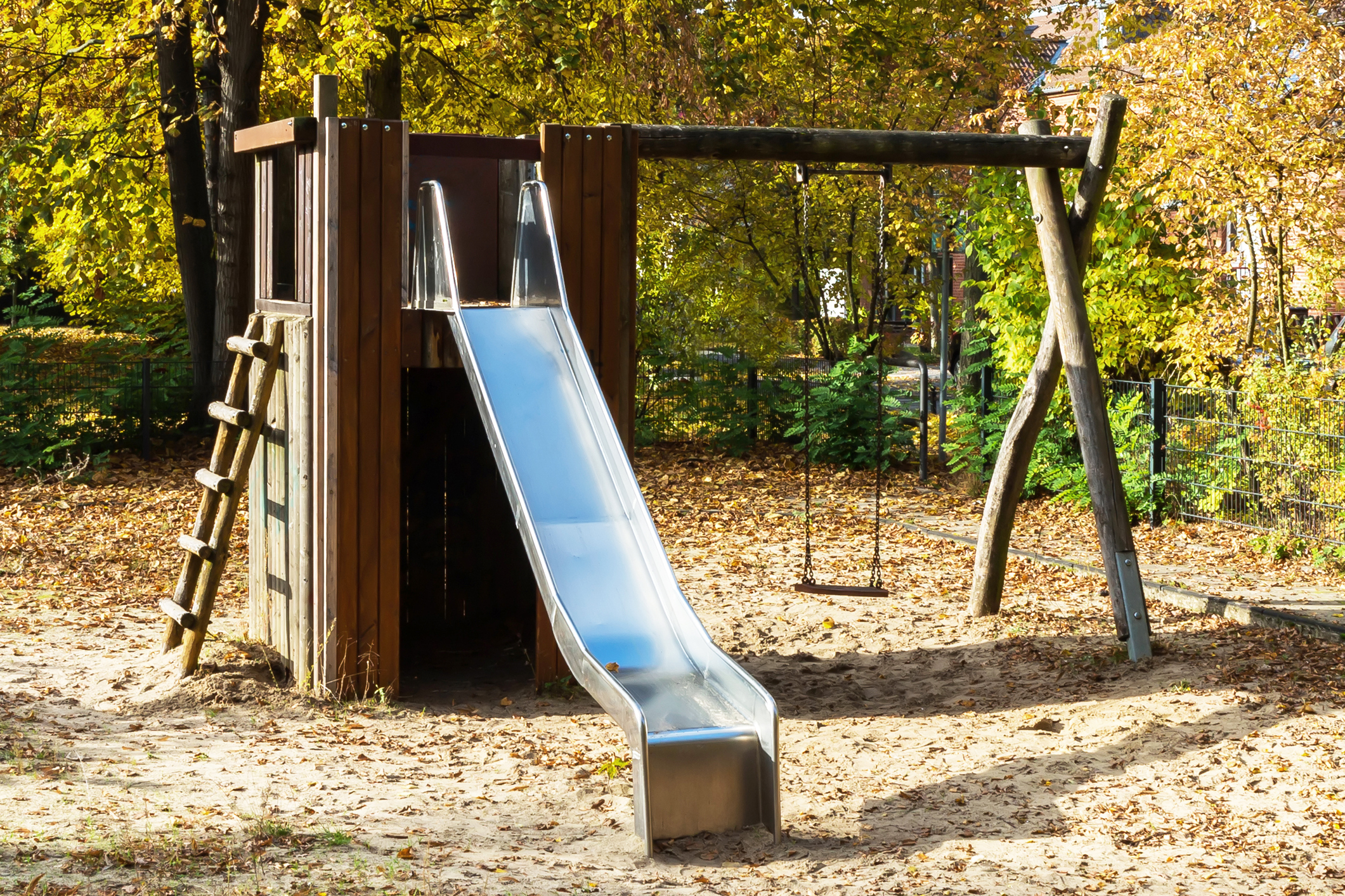 Playground with a slide, swing set, and climbing ladder amidst trees and fallen leaves