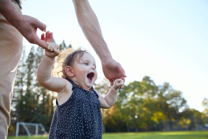 Toddler walking with adult&#x27;s support, expressing joy, outdoors in a park setting