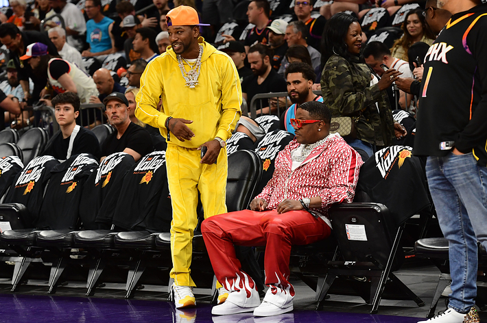 Two men standing courtside at a basketball game, one in a yellow outfit and the other in a red patterned jacket