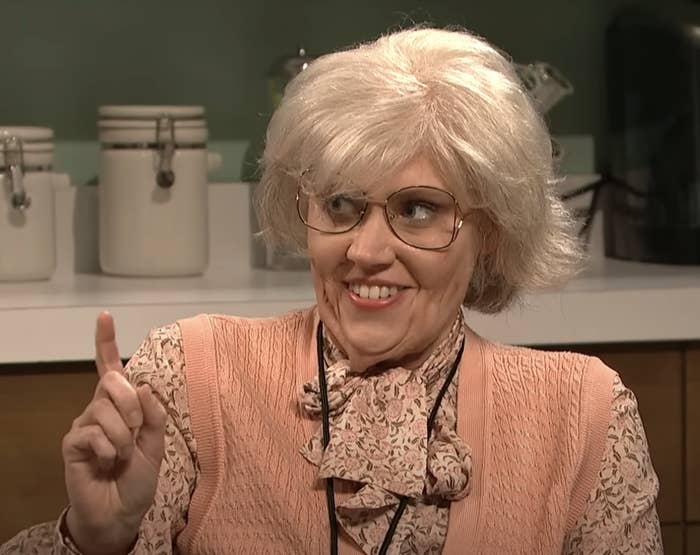 Person dressed as a quirky older lady with a wig and glasses, raising a finger as if to make a point