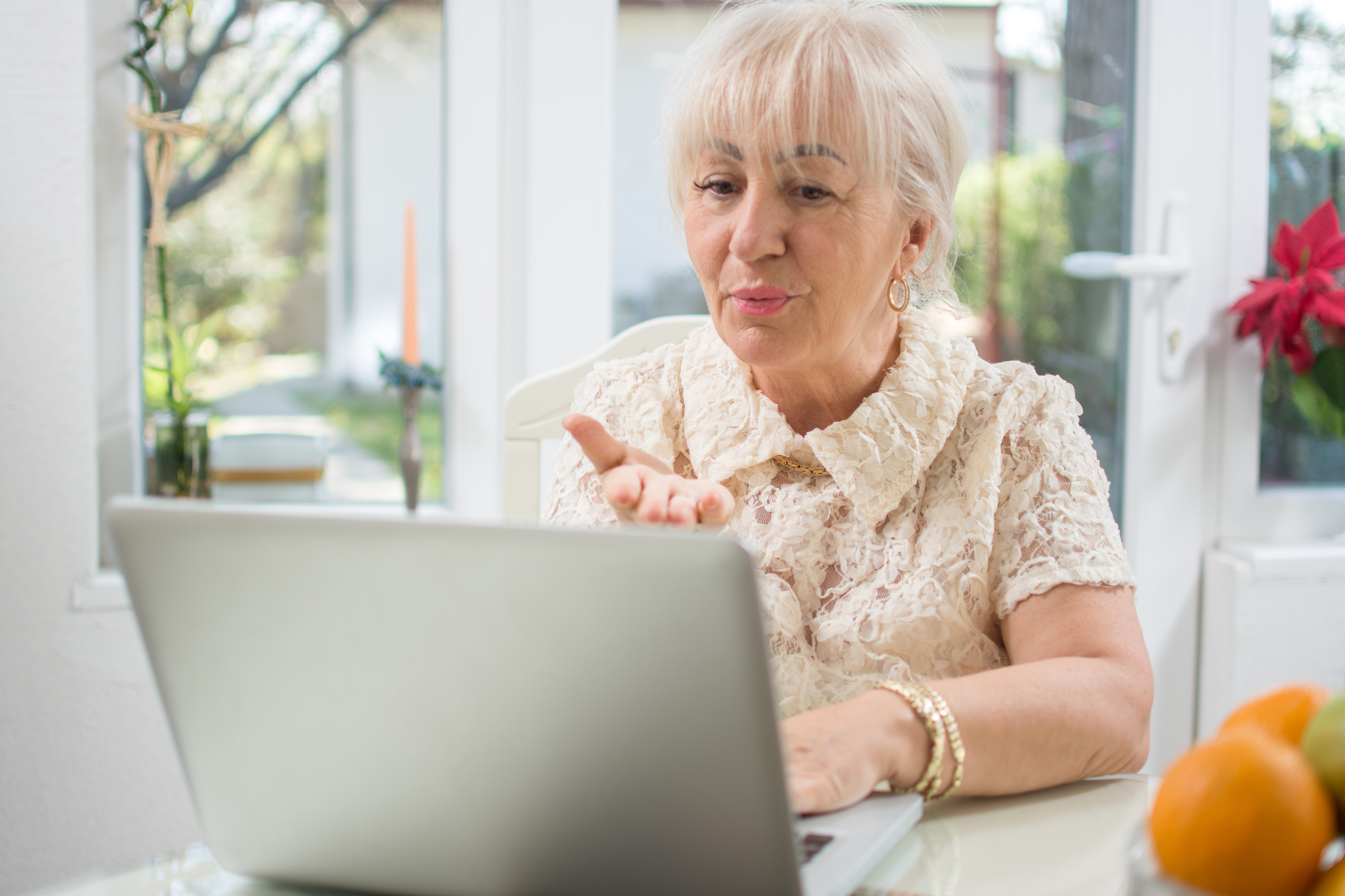 Elderly woman gesturing during a laptop video call, with fruit bowl nearby