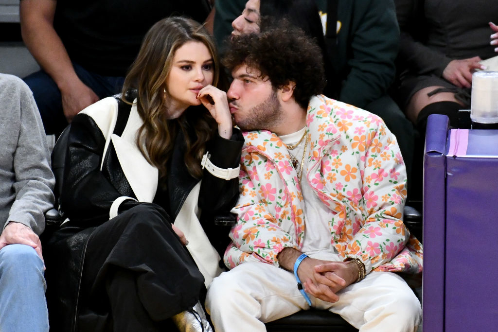 Selena and Benny sitting courtside, Benny leaning over to kiss her hand