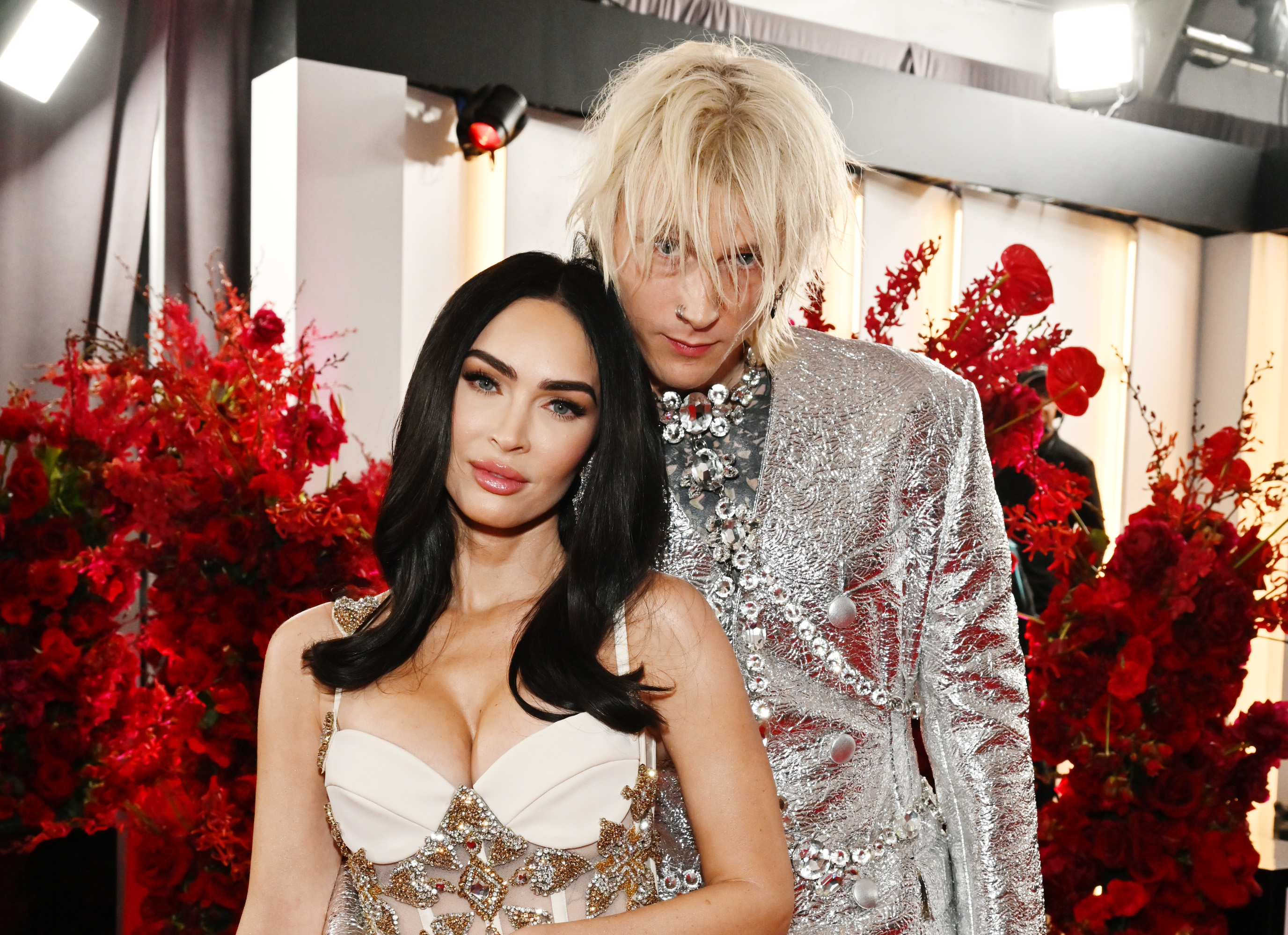 Megan in a bejeweled gown with deep neckline, posing with MGK in a glittery suit at a media event