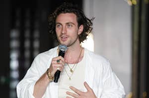 Person speaking into a microphone wearing a casual white shirt, layered necklaces, and wrist accessories