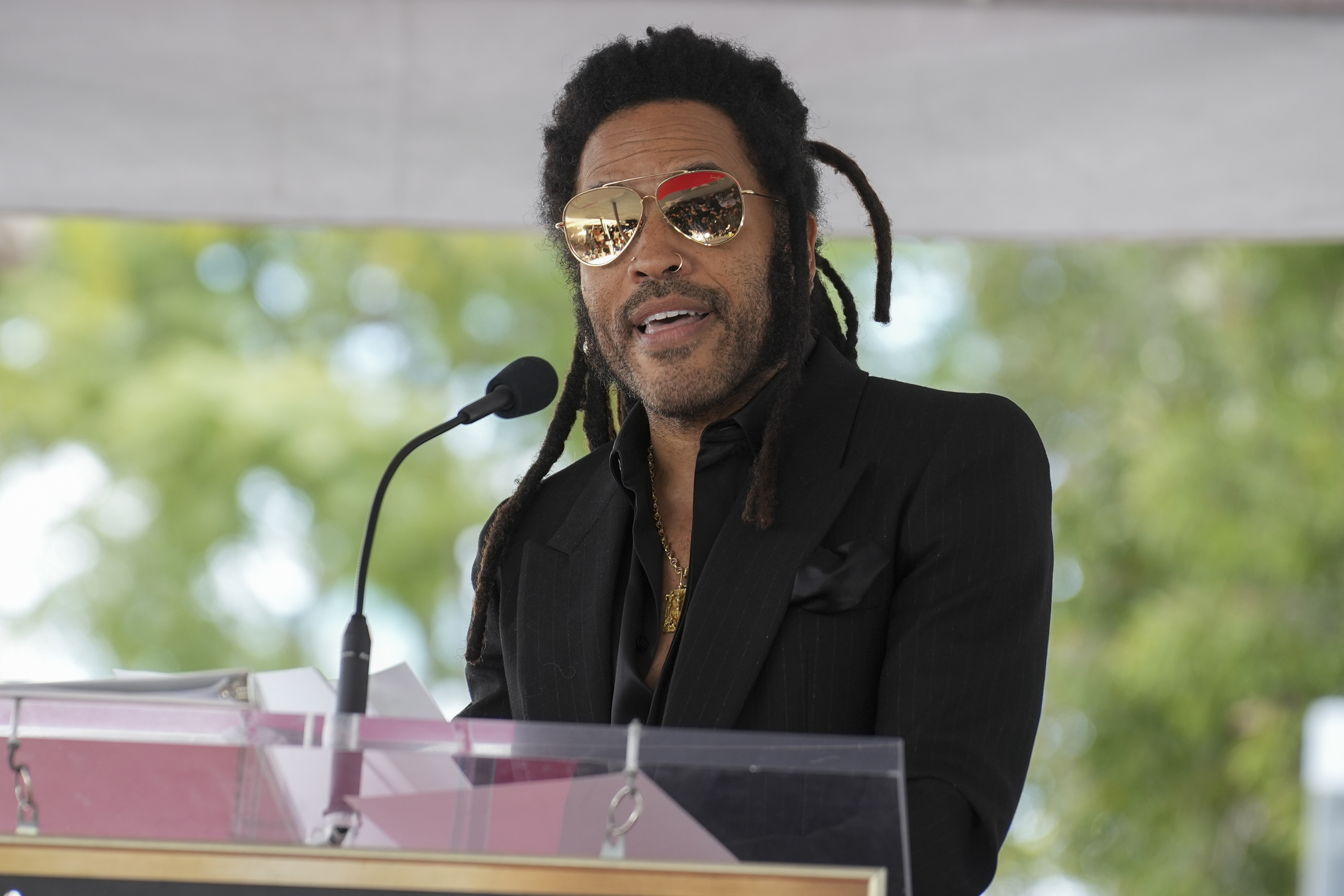 Lenny Kravitz wearing a suit and aviator sunglasses, speaks at a podium