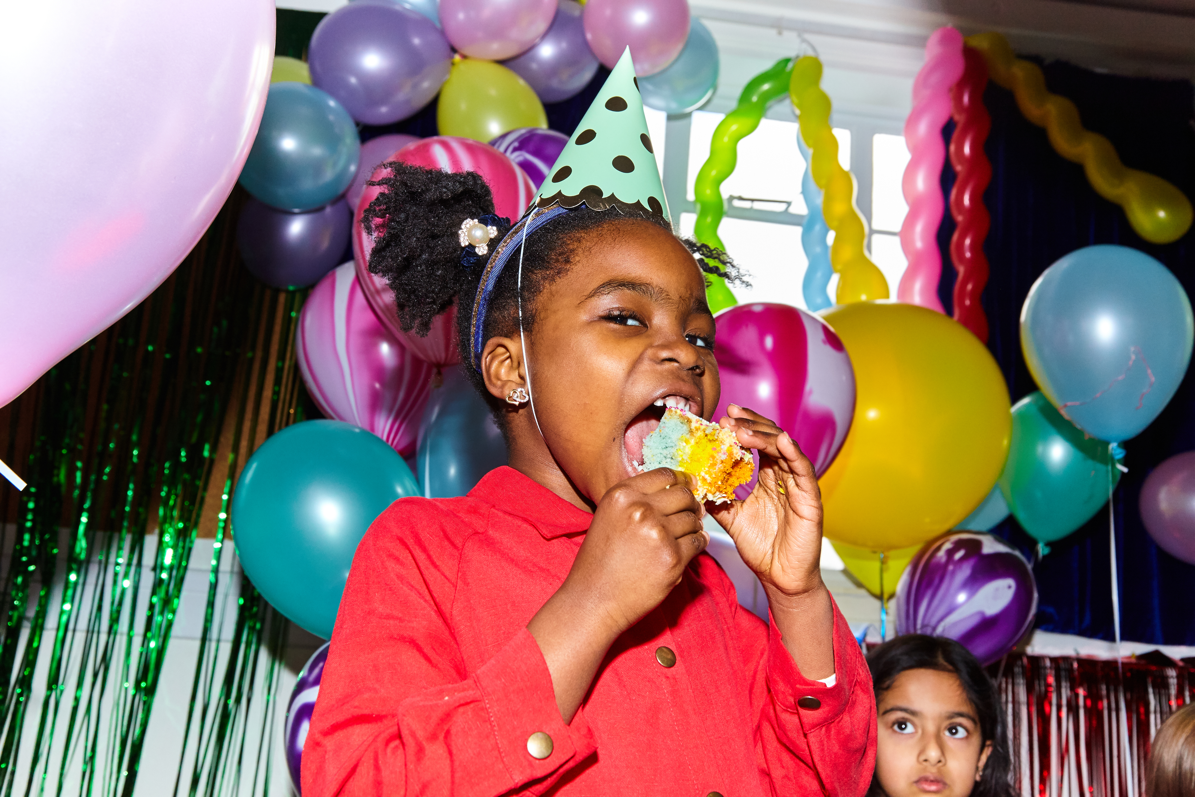 A girl in a party hat enjoys cake at a festive birthday celebration with balloons in the background