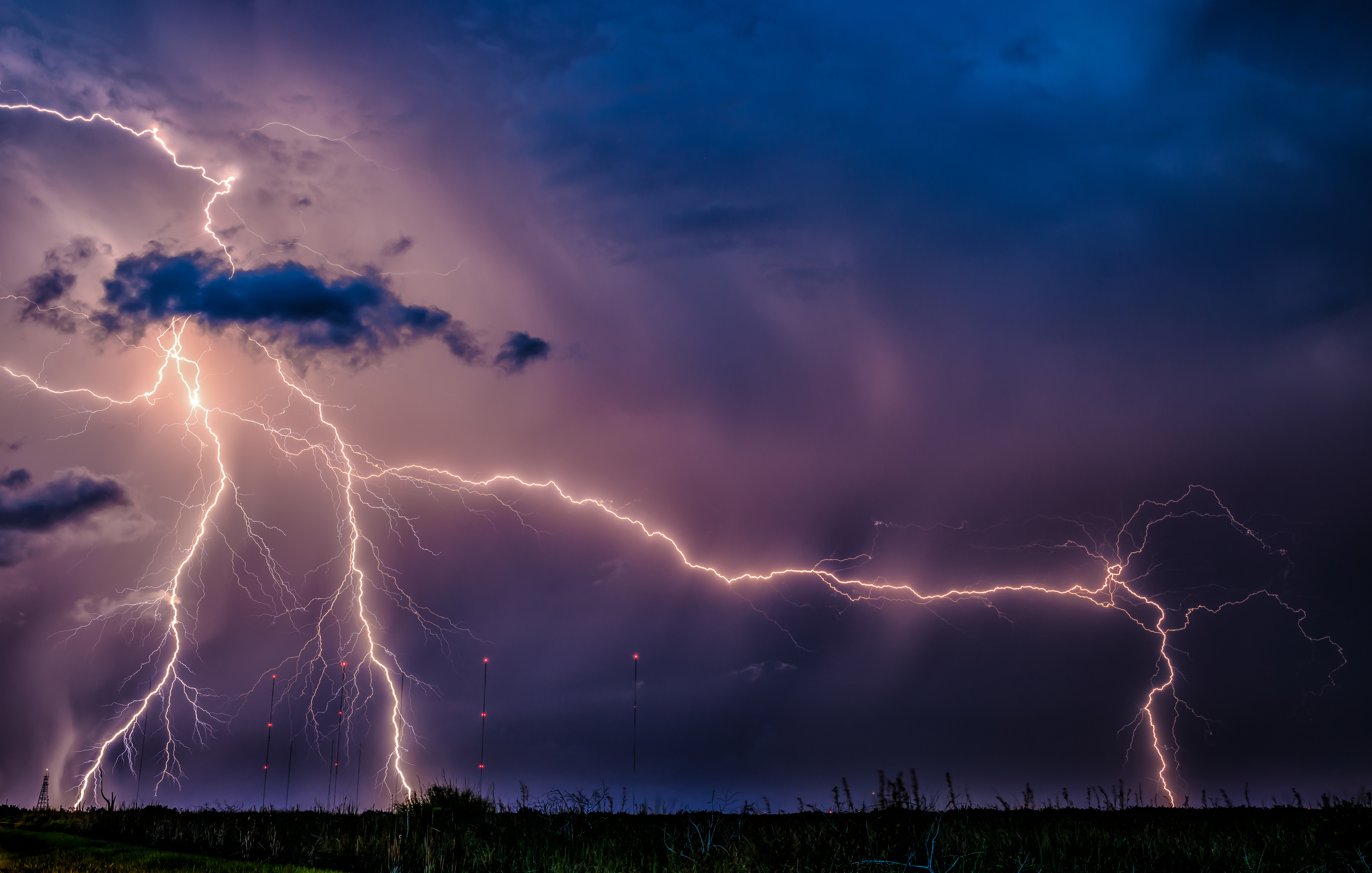 Lightning strikes with visible bolts against a stormy sky, over a field with distant towers