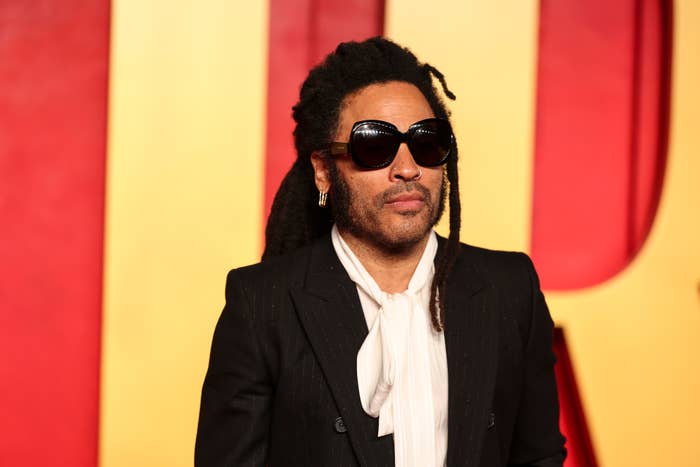 Lenny Kravitz in a black suit and sunglasses, standing in front of a backdrop with logos