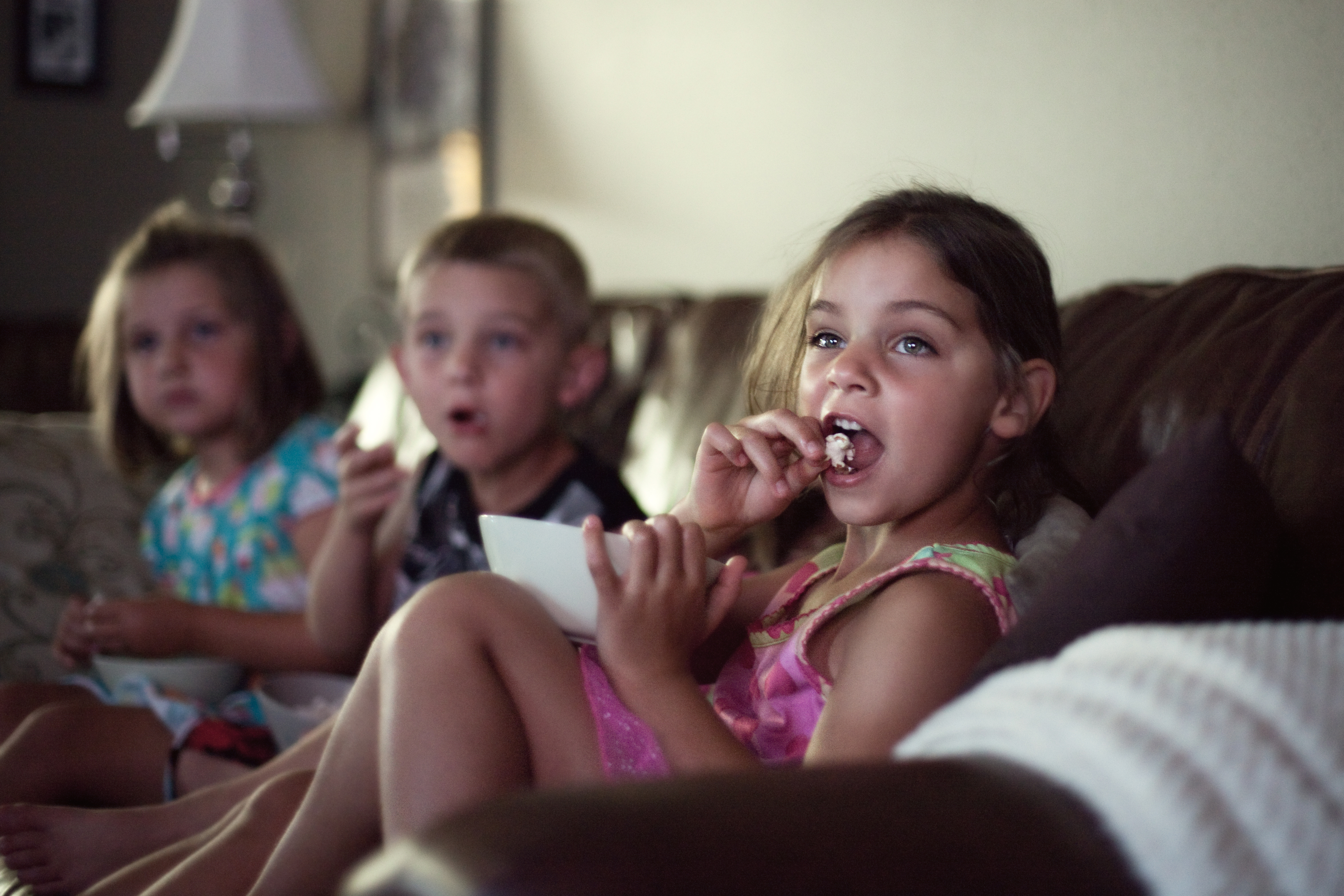 Three children sitting on a couch, engrossed in watching TV, one eating a snack