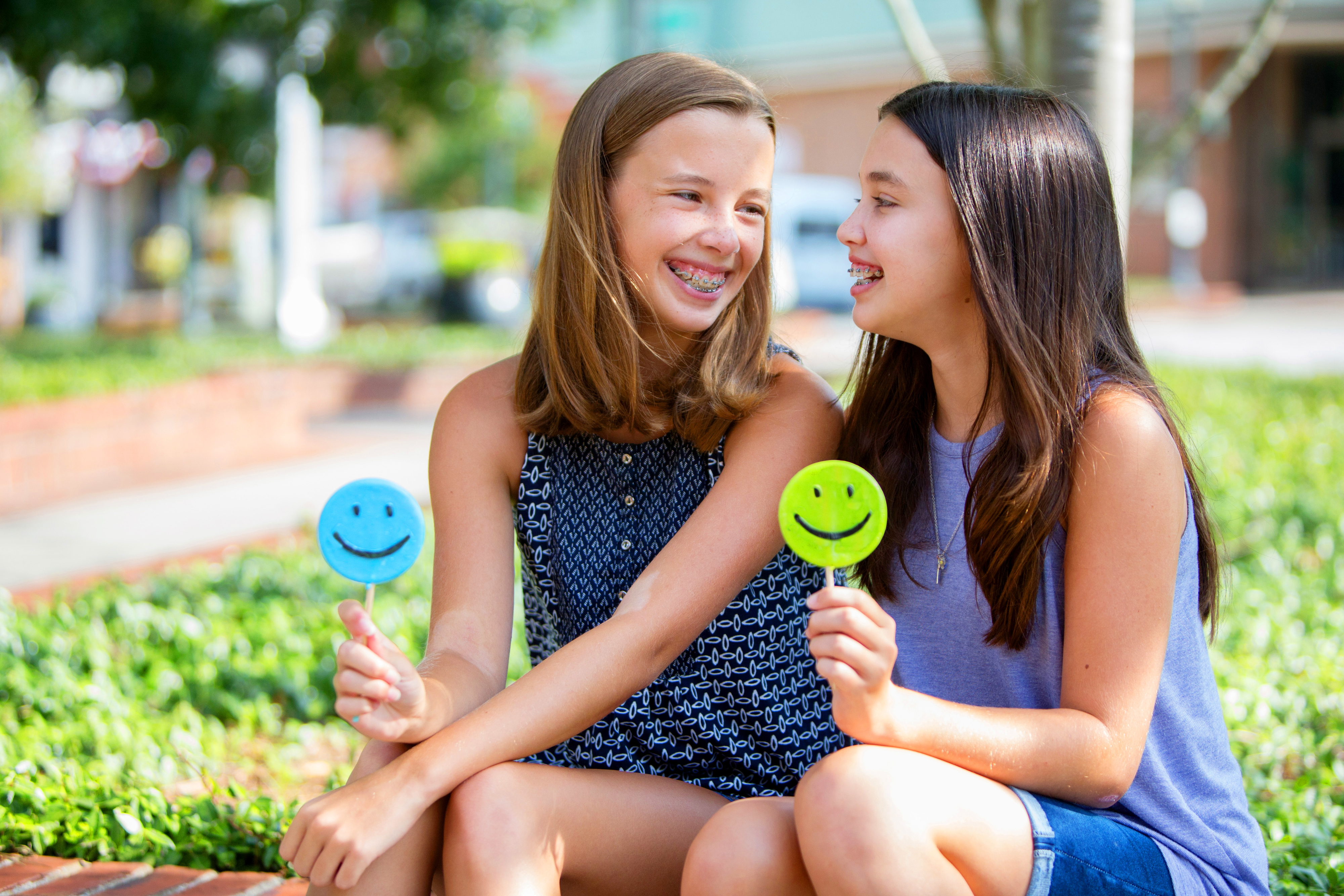 Two girls sitting outside, smiling, holding lollipop smiley faces