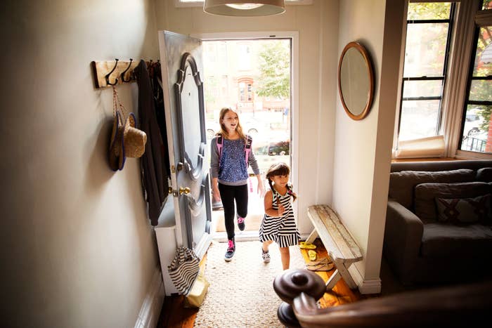 Two children entering a house, one wearing striped attire, with bags and coats hanging by the door