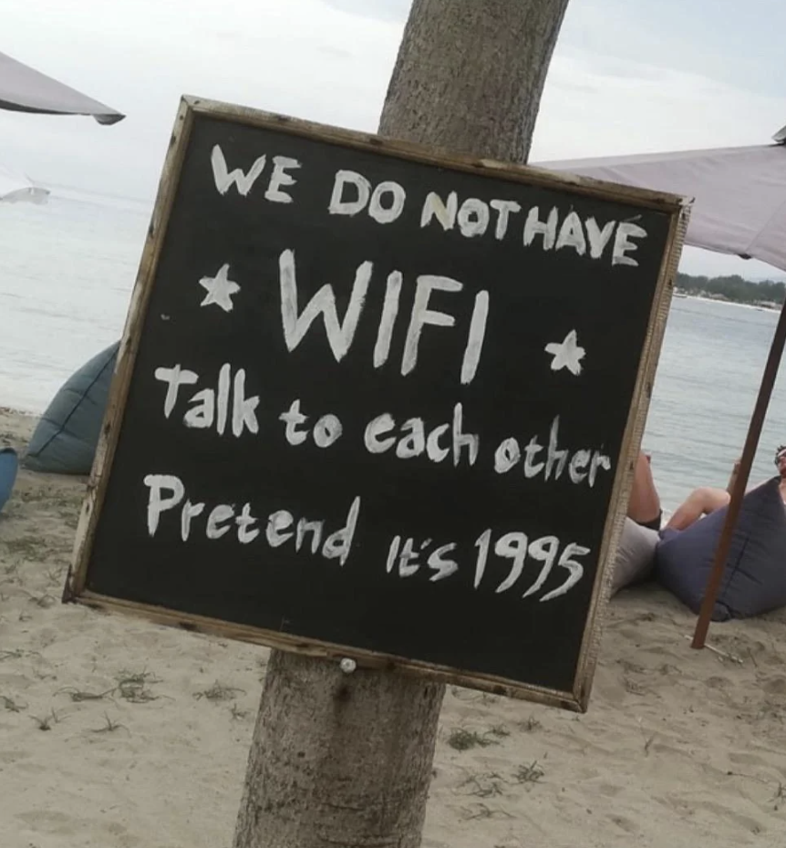 Chalkboard sign on a beach reads &quot;WE DO NOT HAVE WIFI talk to each other Pretend it&#x27;s 1995.&quot;
