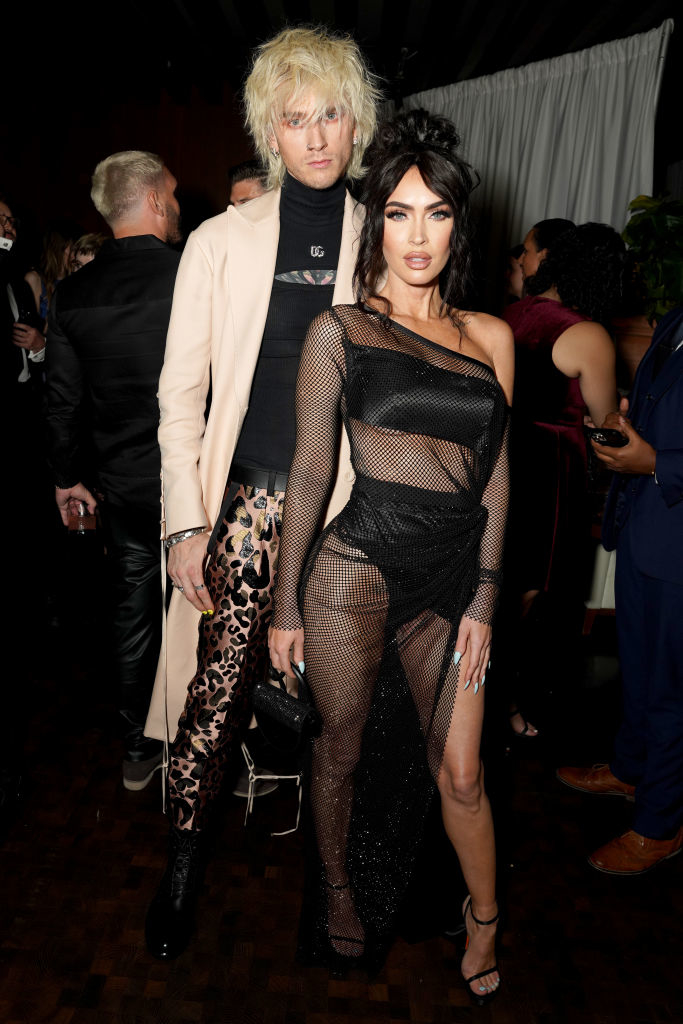 MGK and Megan posing, one in a beige jacket and leopard print pants, the other in a sheer black outfit with mesh details