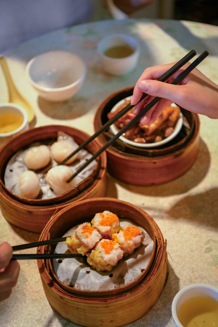 Person using chopsticks to pick dim sum from a bamboo steamer at a table with tea