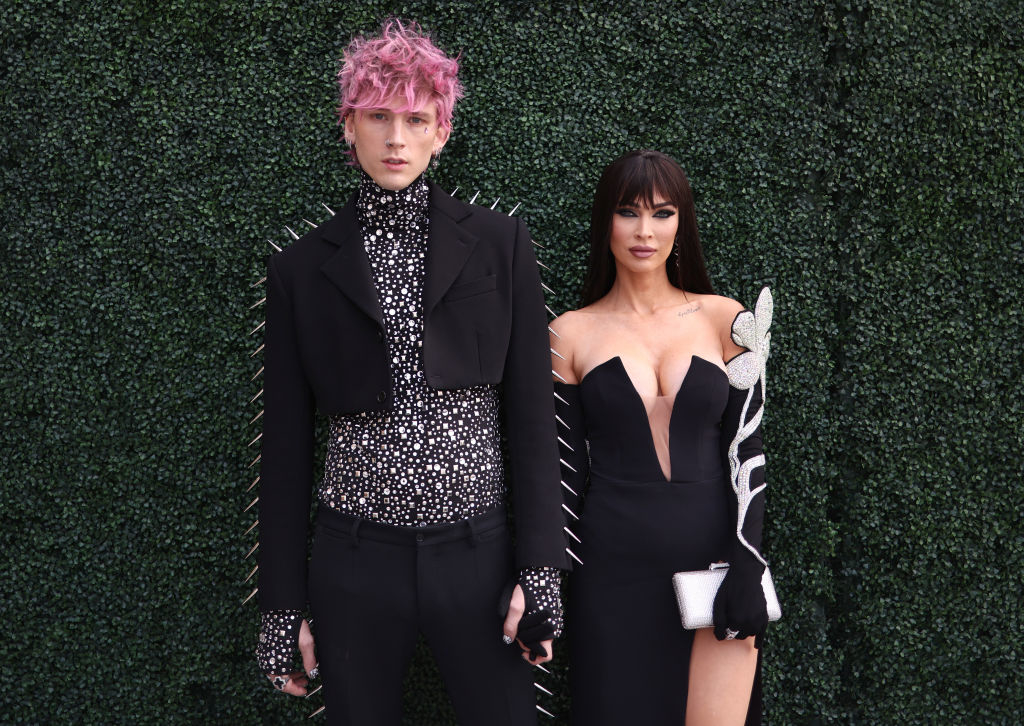 MGK and Megan at an event, one in a studded jacket and patterned shirt, the other in a black dress with white accents