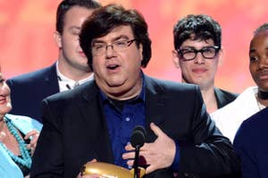 Man in glasses and blue shirt holding award, speaking at a microphone with people in background