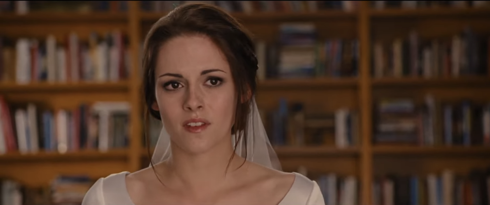 Bella Swan in a simple wedding dress with a sheer veil, standing in a room with bookshelves