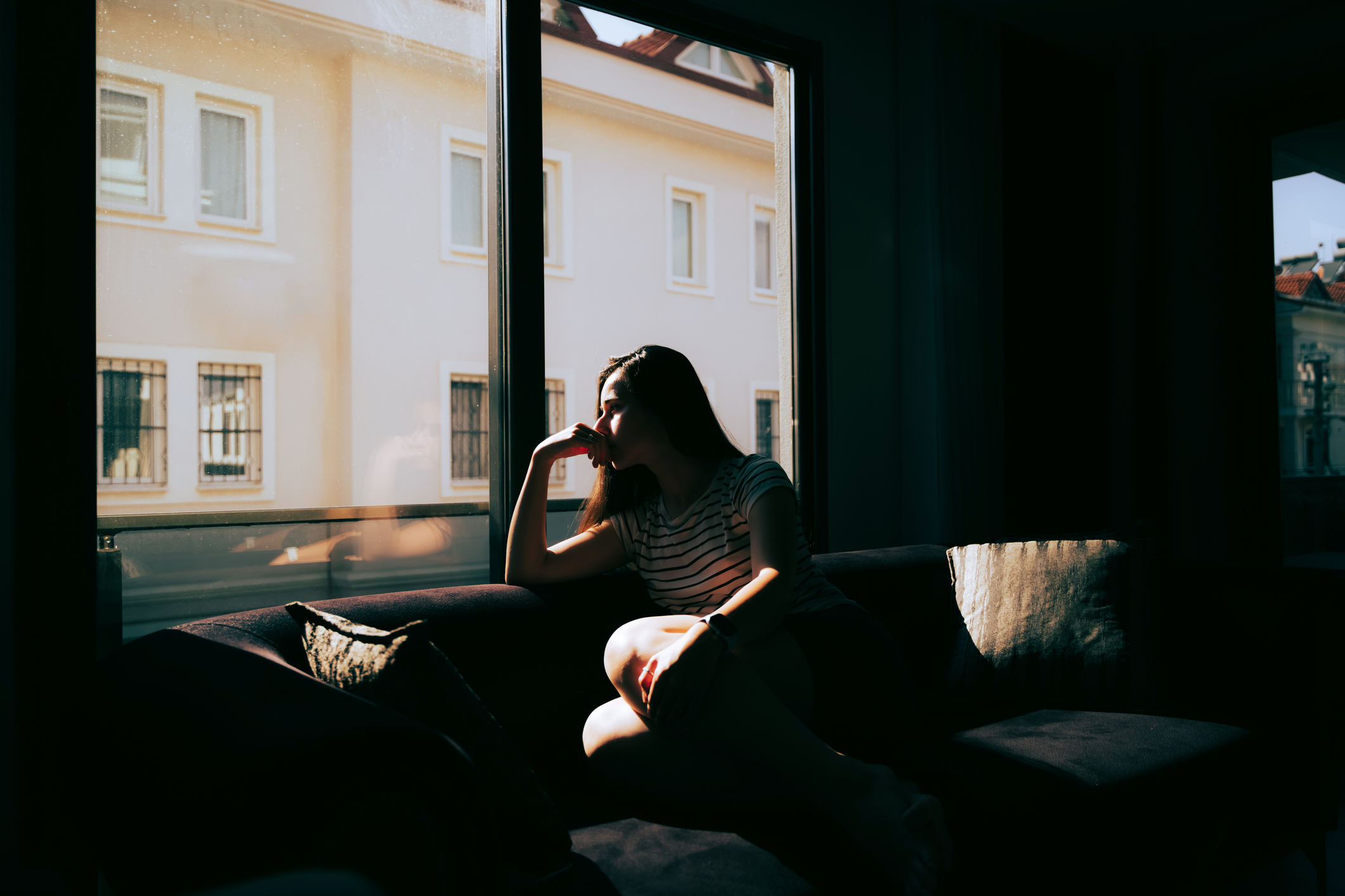 Person sitting by a window in a contemplative pose, sunlight casting shadows indoors