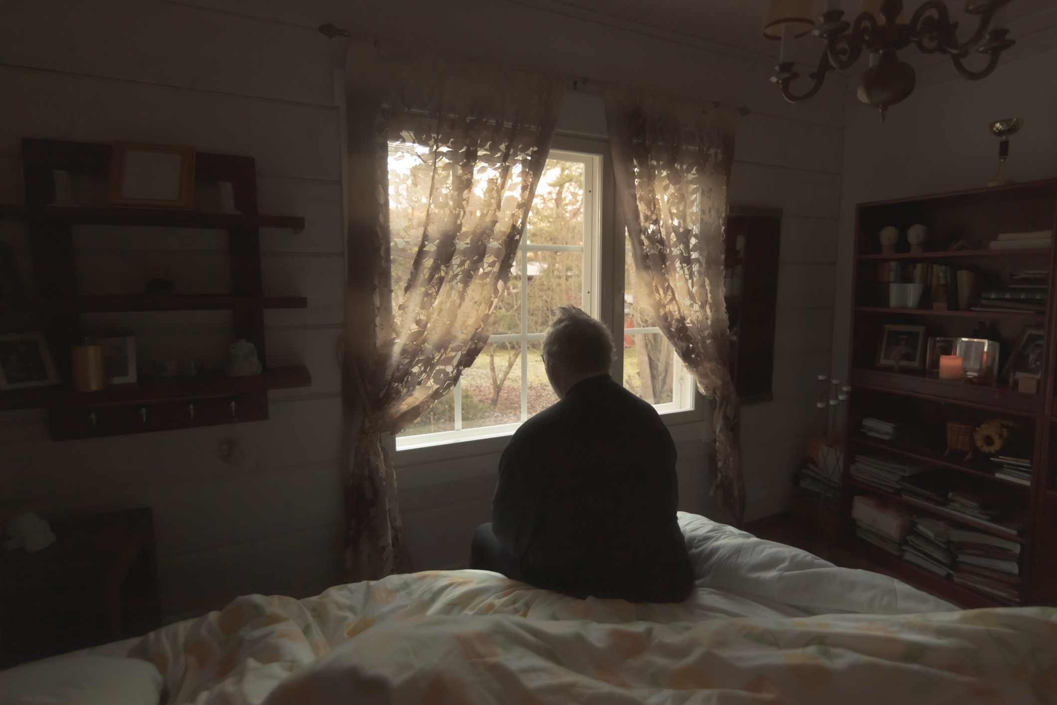 Person sitting on a bed looking out of a window in a room with sheer curtains and a chandelier