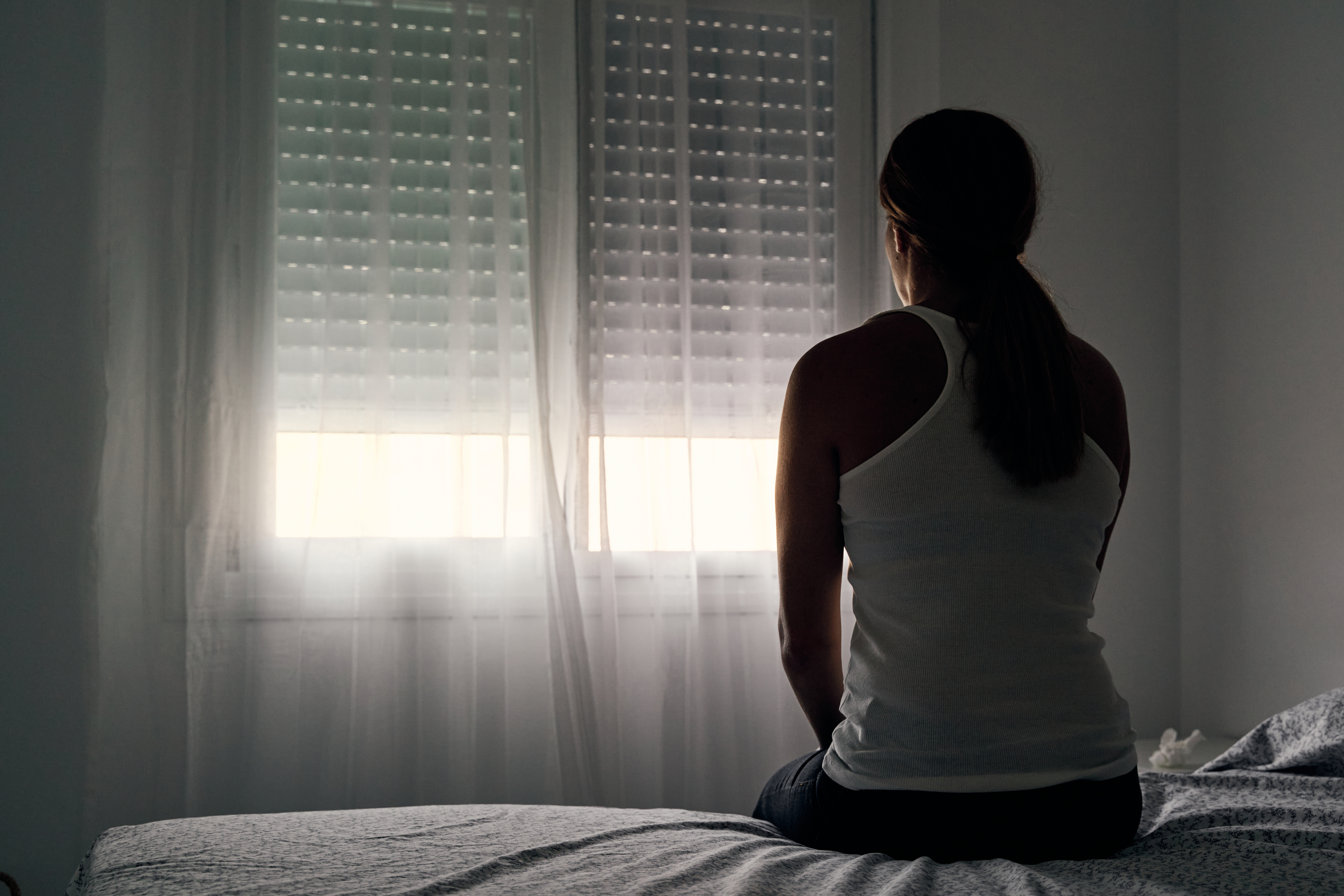 Woman sitting on a bed facing a window with closed shutters, room appears dimly lit