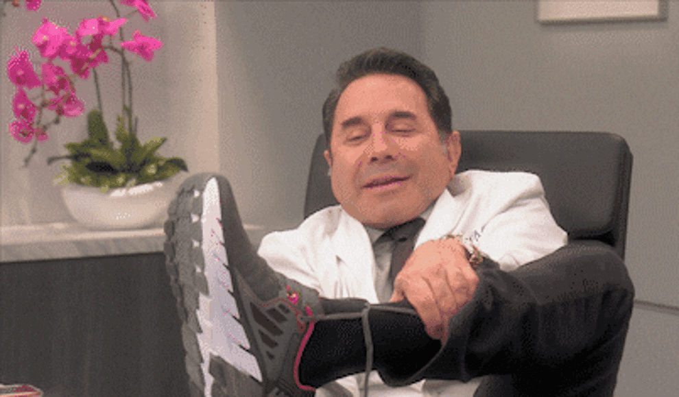 Dr. Paul Nassif, lifting a large sneaker near his face in an office setting