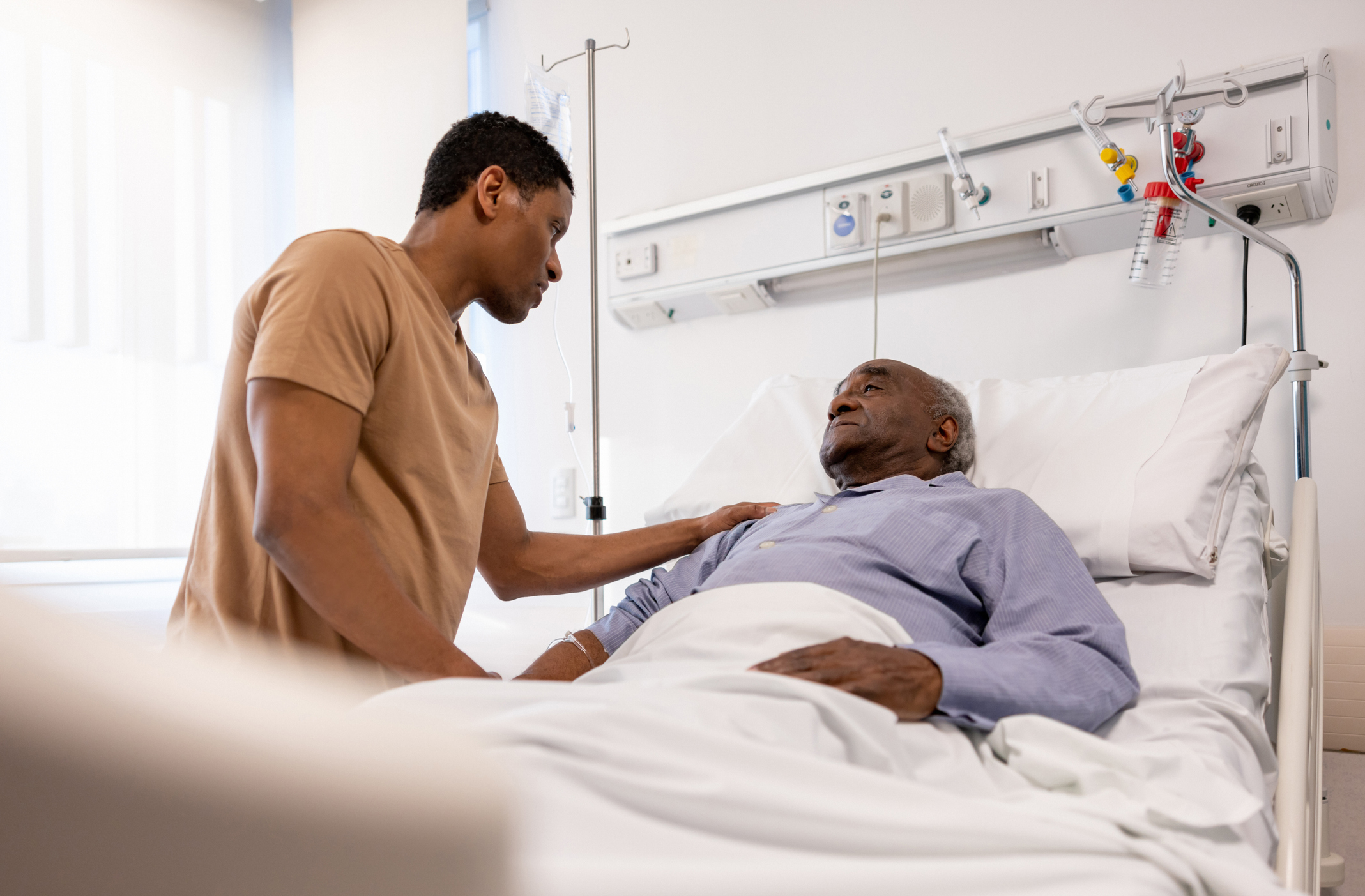 A younger person comforting an older person lying in a hospital bed