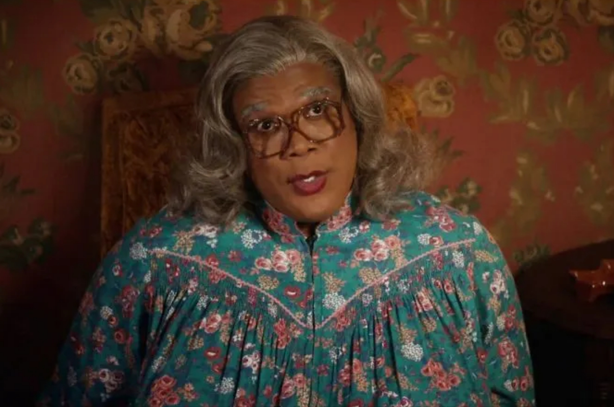 Tyler Perry as Madea, wearing a floral blouse, looking sternly forward from a floral wallpaper background