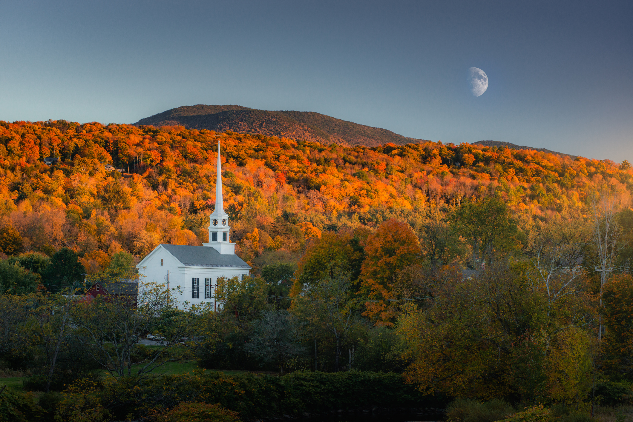 A church with a tall spire in front of a forested hill with fall foliage under a sky with a visible half moon