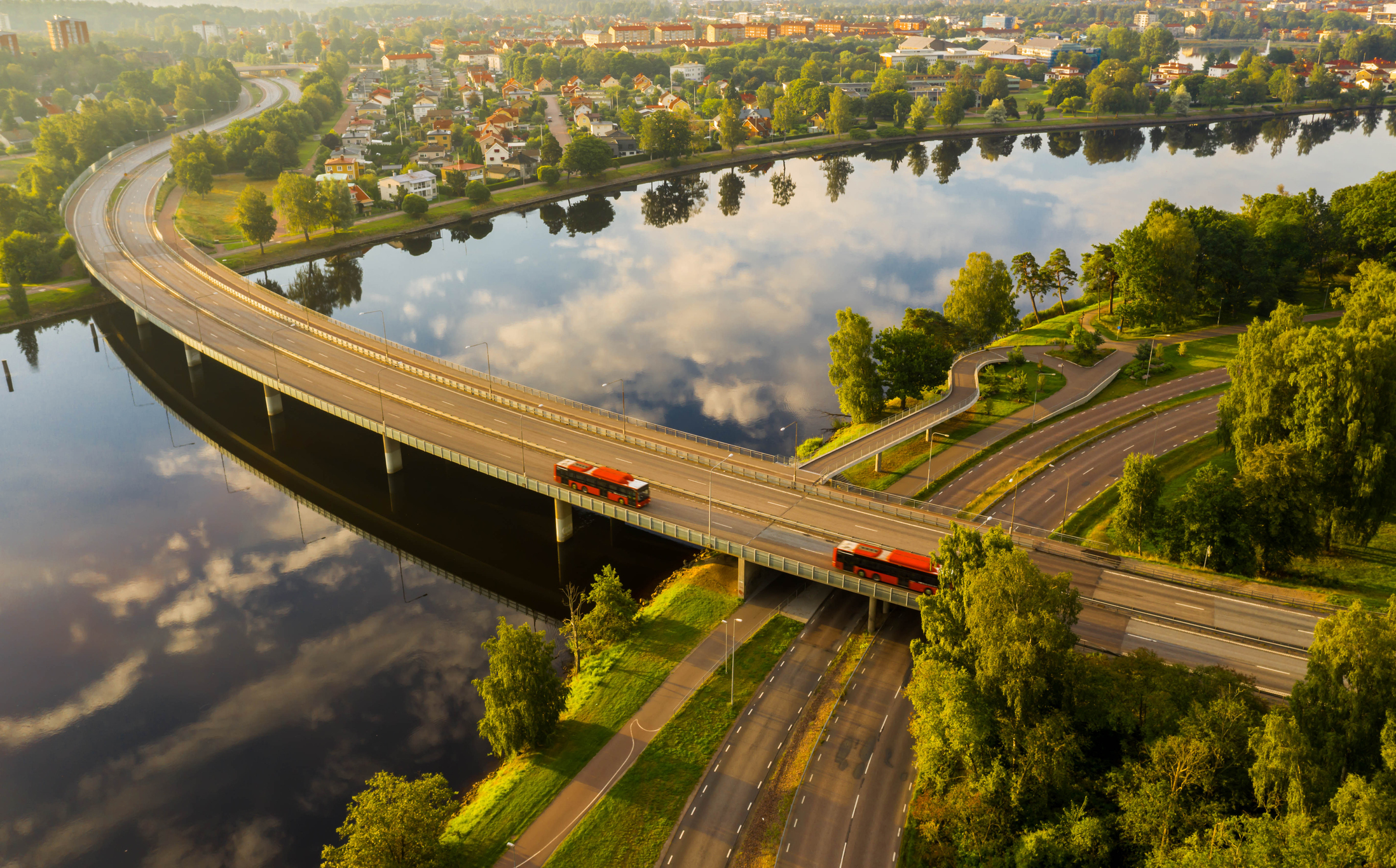 Aerial view of a bridge over a river with a red train crossing, adjacent roads, and surrounding greenery