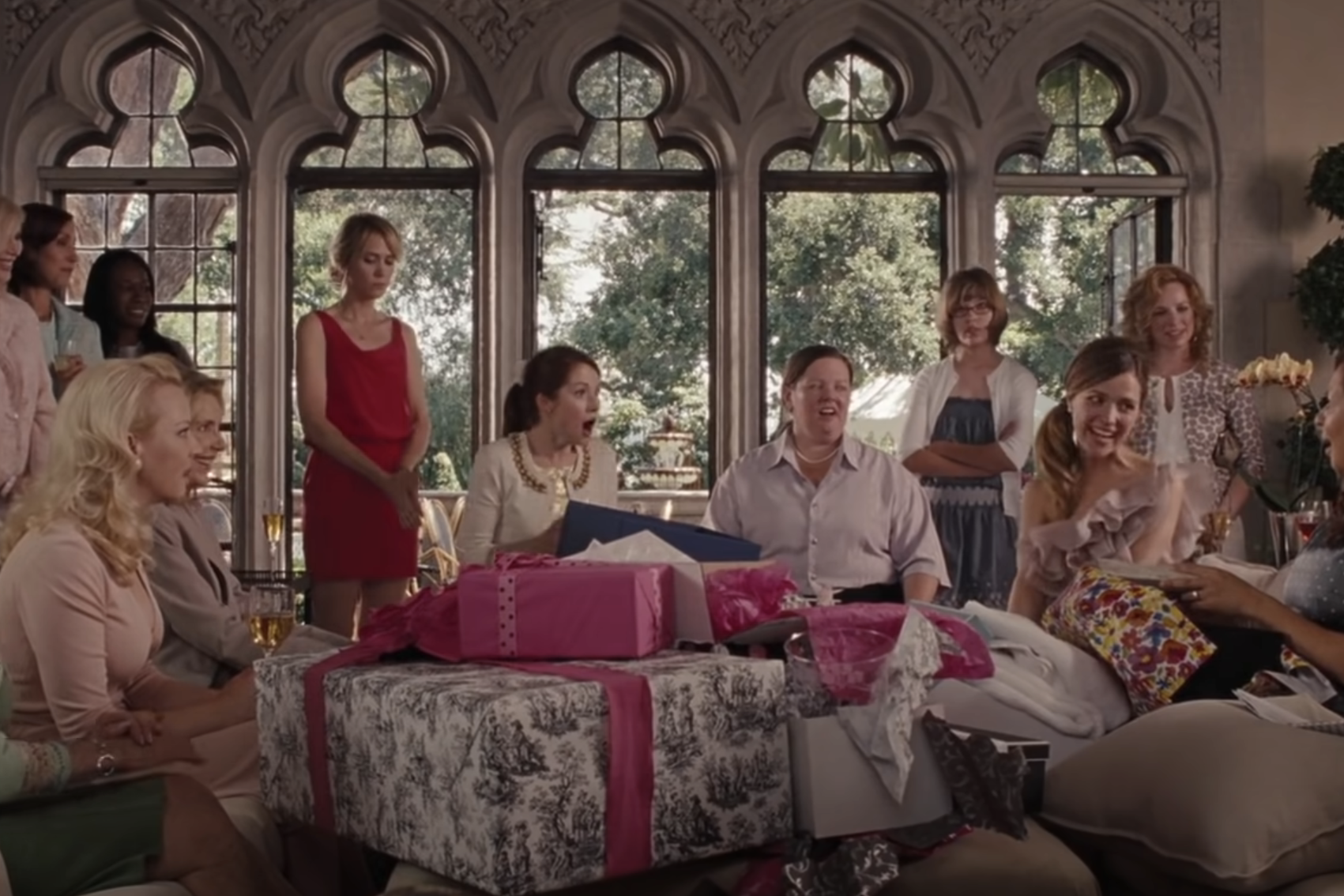 Group of women at a gift-giving gathering in a stylish living room with ornate windows, piles of wrapped presents, and smiles all around