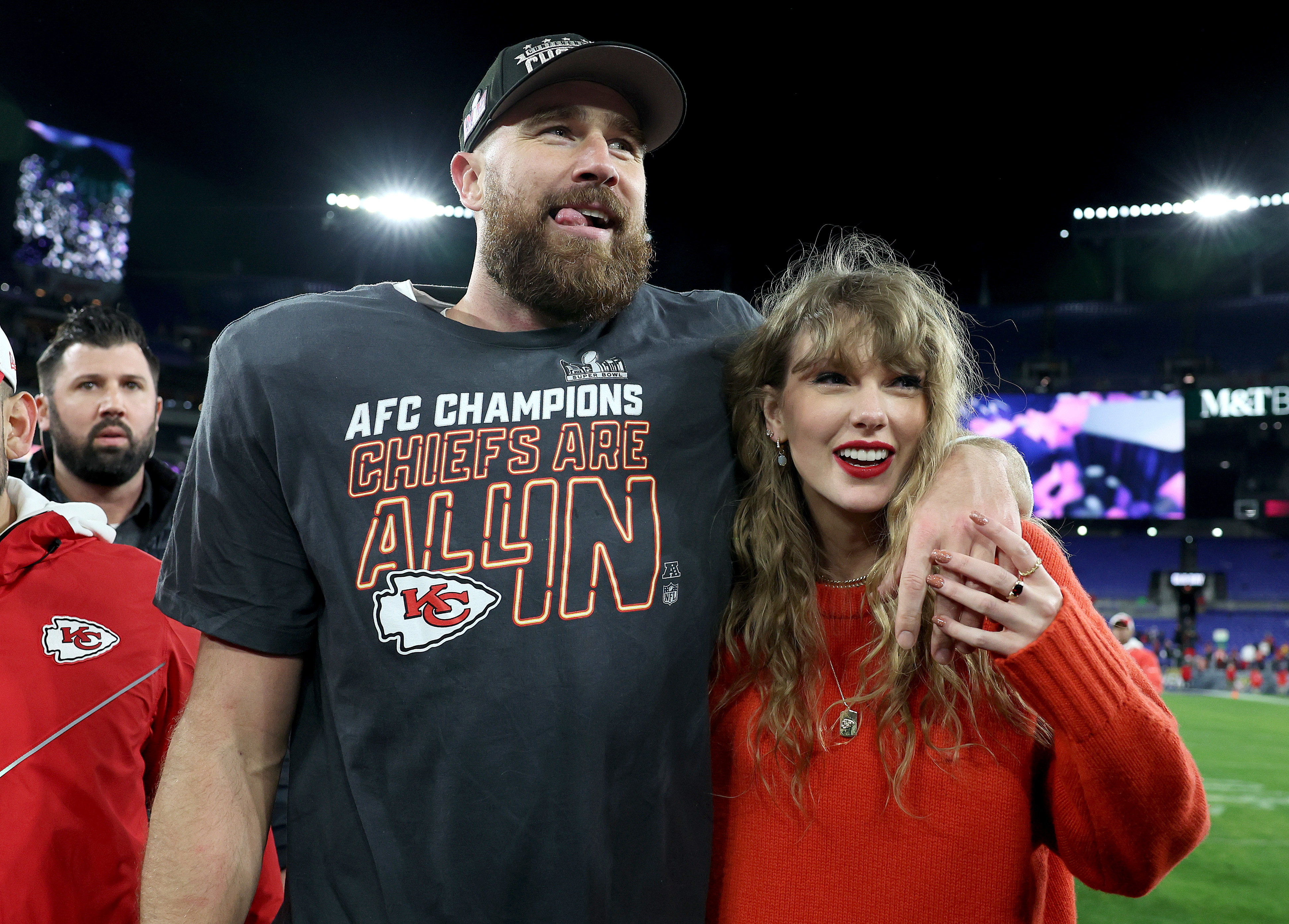 Travis Kelce and Taylor Swift smiling together at a sports event, Travis wearing a Chiefs t-shirt