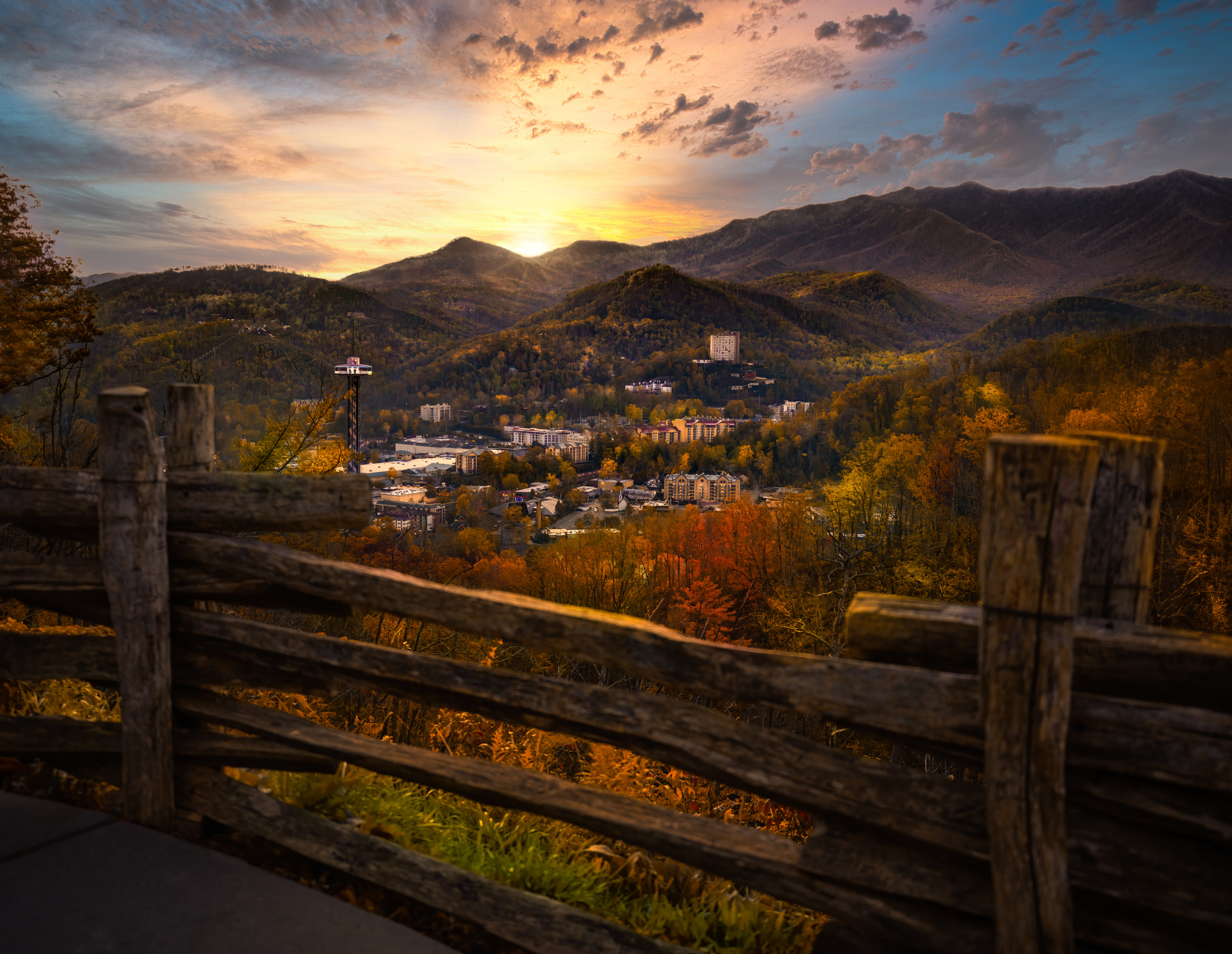 View of a town nestled in autumnal mountains at sunset, seen from behind a rustic wooden fence