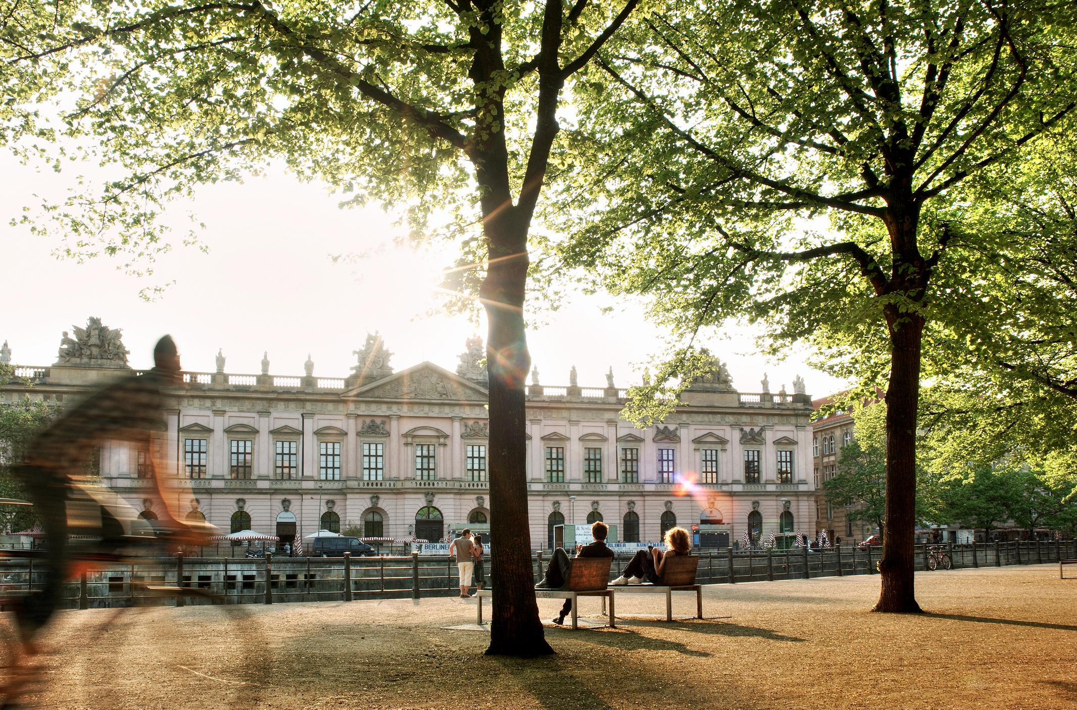 People relax by benches near trees with a classical building in the background. A blurred figure is in motion