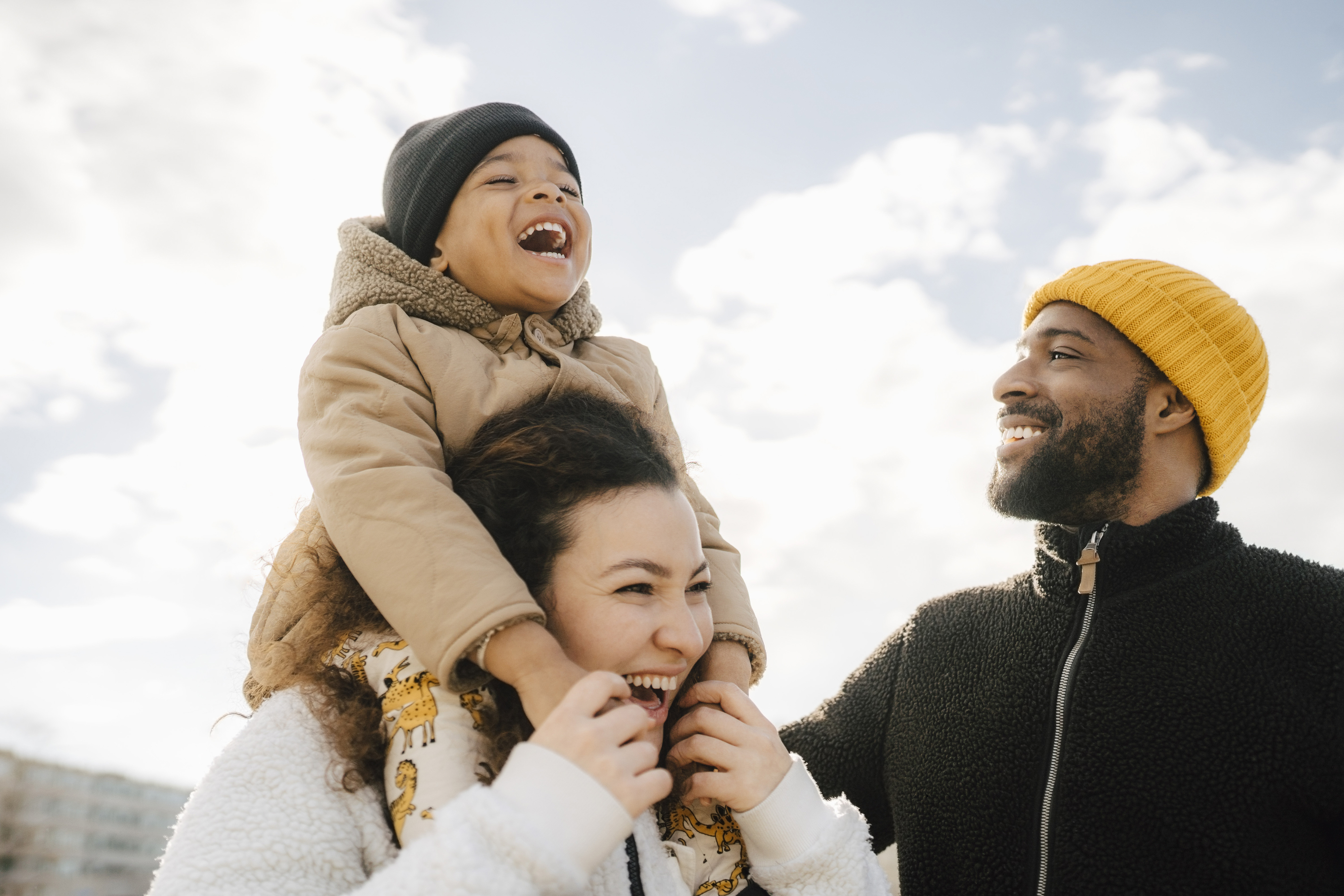 Child on woman&#x27;s shoulders, man beside them smiling, outdoors, casual attire, family moment