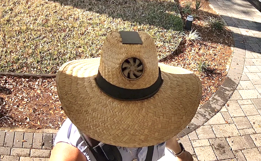 Person with a large-brimmed straw hat with a fan on the crown, in a sunny outdoor setting