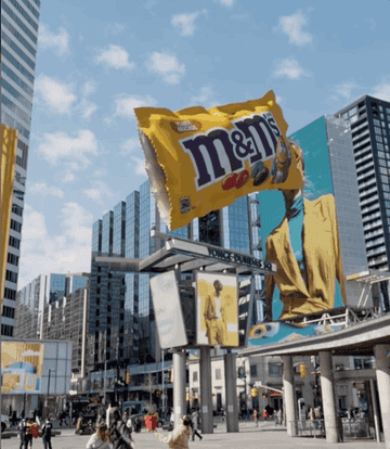Giant M&amp;amp;M&#x27;s candy bag above a cityscape with pedestrians and modern buildings