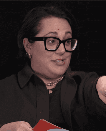 Person gesturing with left hand, wearing glasses and a black top, with a watch on left wrist