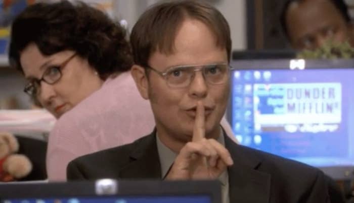 Dwight from &quot;The Office&quot; gestures for silence with a finger over his lips, Phyllis in background. They&#x27;re in an office setting
