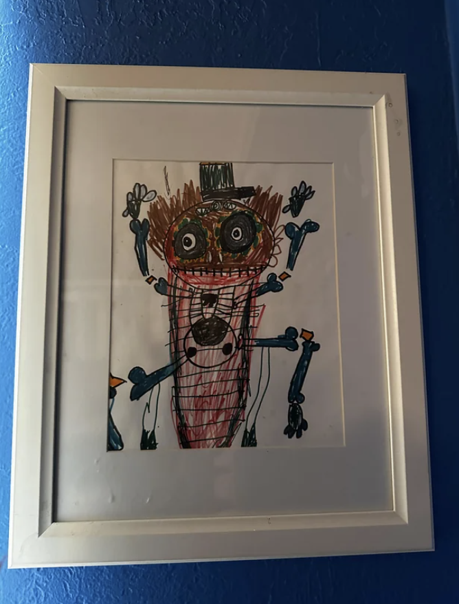 Child&#x27;s drawing of a whimsical figure with a top hat, framed and hung on a blue wall