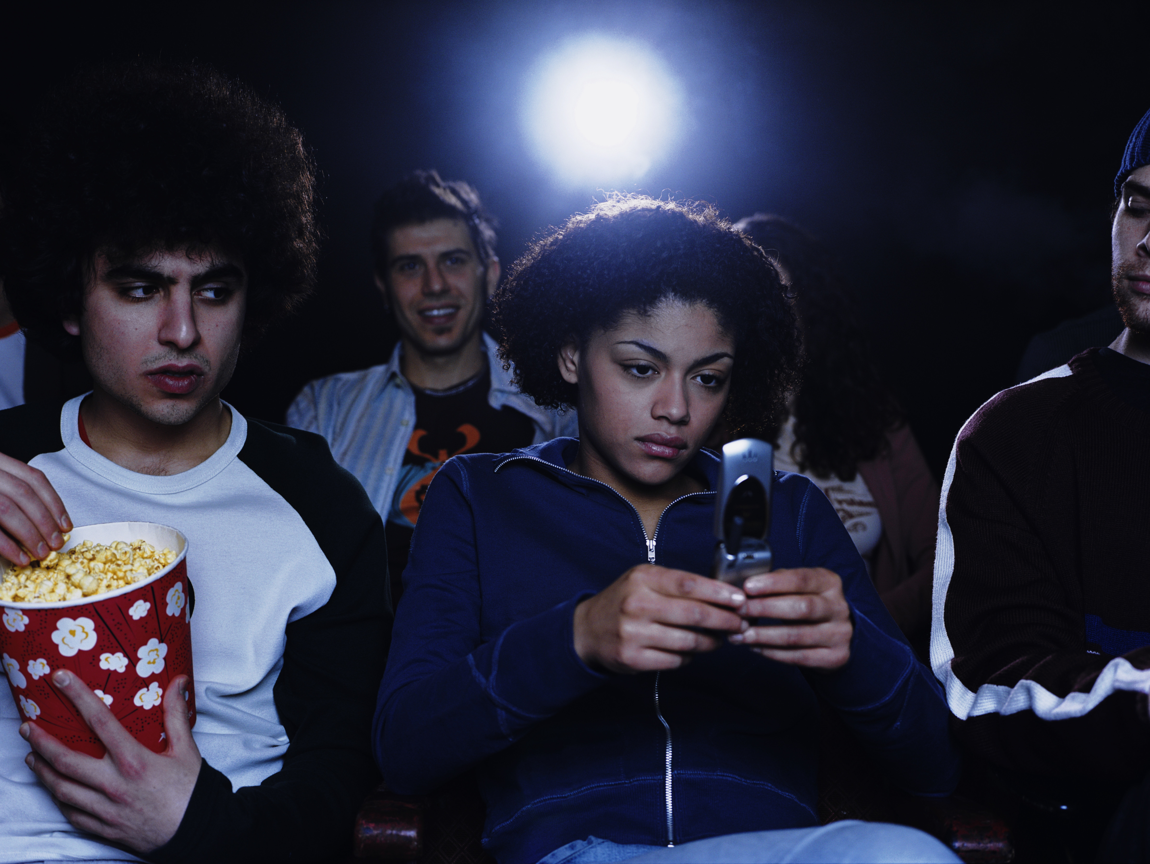 People in a dark cinema; one is focused on their phone screen, others watch the film, one with popcorn