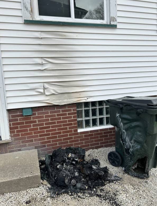 Melted siding on a house above a pile of burnt debris next to a trash bin